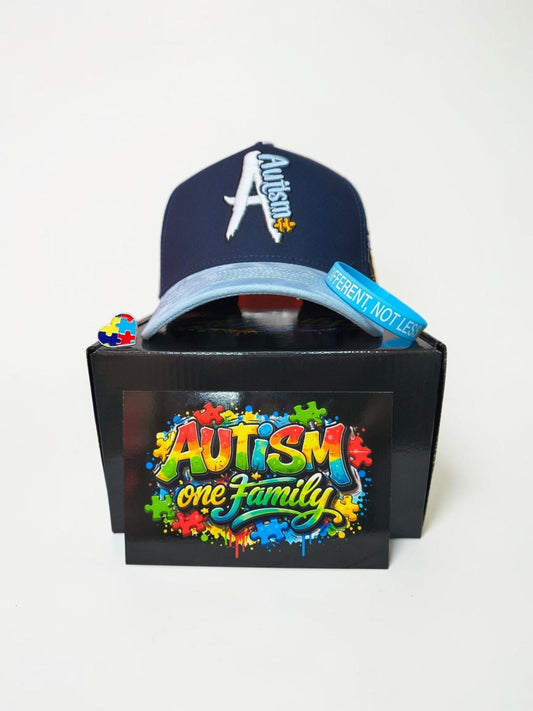 A navy blue baseball cap with 'Autism' written in white and yellow, displayed on top of a black box featuring a colorful 'Autism one Family' graphic. 